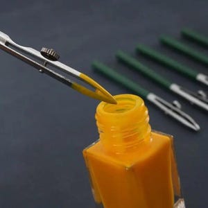 May include: A silver metal ruling pen with a yellow-coated tip is positioned above an open square glass bottle filled with bright yellow ink. Several additional ruling pens with dark green handles are arranged in the background.