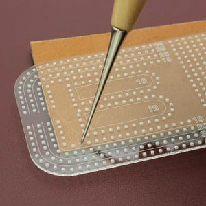 Puede incluir: Una plantilla de acrílico transparente para costura de cuero con patrones de agujeros precortados y marcas numéricas, incluyendo 10, 12, 15, 25, 35 y 45, colocada sobre una pieza de cuero beige con una lezna de metal con mango de madera.