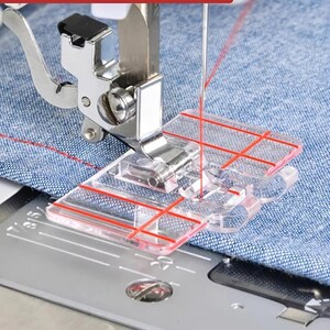 May include: A clear plastic sewing machine presser foot with red alignment grid lines attached to a metal shank. The foot is positioned over blue fabric with a red thread line, resting on a metal sewing machine plate with visible markings.