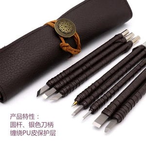 May include: A set of carving tools with silver metal blades and brown leather-wrapped handles, accompanied by a dark brown leather roll case secured with a tan cord and a circular brass-toned decorative button. Text reads: 产品特性：圆杆、银色刀柄、缠绕PU皮保护层.