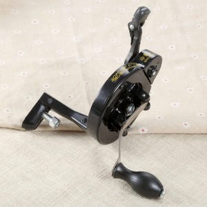 May include: A black metal hand-crank sewing machine attachment featuring a circular gear housing with gold decorative scrollwork, a silver-toned metal handle with a black knob, and a mounting bracket with a silver-toned metal tightening bolt.