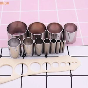 May include: A set of eleven cylindrical metal cutters arranged in descending order of size, accompanied by a piece of off-white rolled dough featuring eleven circular cutouts that correspond to the diameters of the metal tools.