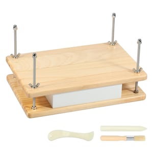 May include: A light-coloured wooden bookbinding press featuring two rectangular boards held together by four threaded metal rods with wing nuts. A white rectangular block is positioned between the boards. Includes a bone folder and a brush.