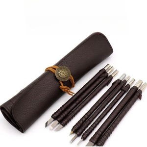 Könnte beinhalten: Ein Set aus sechs Schnitzwerkzeugen mit dunkelbraun umwickelten Griffen und Metallspitzen, begleitet von einer passenden, dunkelbraunen, texturierten Rolltasche, die mit einer beigen Lederschnur und einem dekorativen, antik-bronzenen Knopf verschlossen wird.