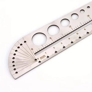 以下が含まれることがあります： 円形の切り抜きがある曲線状の分度器エッジと、直線の定規エッジを備えた銀色の金属製製図ツール。ツールにはミリメートル単位の目盛りが刻印されており、直線エッジに沿って1から6までの数字が確認できます。
