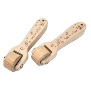 Pode incluir: Dois rolos de costura de madeira clara com uma roda cilíndrica numa extremidade. Cada cabo apresenta o texto gravado "Seam roller" acompanhado por um pequeno desenho de borboleta e flores. As ferramentas têm um acabamento liso e ferragens metálicas.