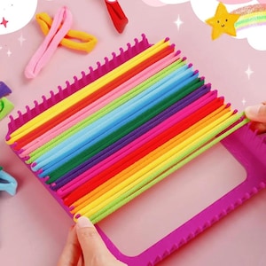Peut inclure: Un cadre de métier à tisser carré en plastique violet avec des rangées de picots sur deux côtés opposés. Des bandes élastiques de couleurs vives, notamment rose, jaune, vert, bleu, violet et rouge, sont tendues sur les picots en rayures.