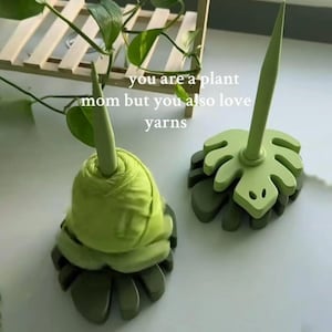 May include: Two green yarn holders shaped like monstera leaves with central spindles. One holder is empty, and the other holds a ball of light green yarn. The text "you are a plant mom but you also love yarns" is visible in the center of the image.