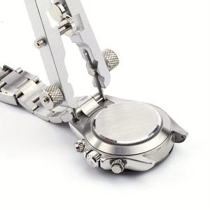 Könnte beinhalten: Ein silberfarbenes Federstegwerkzeug aus Metall mit verstellbaren Rändelschrauben und einem zentralen Rahmen, das so positioniert ist, dass es in die Bandanstöße eines Edelstahl-Armbanduhrgehäuses mit gebürstetem Boden eingreift.