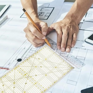 May include: A clear rectangular quilting ruler with yellow and black grid markings, including degree lines and numerical measurements in inches, positioned over architectural floor plan blueprints while being used with a wooden pencil.