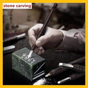 May include: A dark green rectangular stone block being carved with a metal tool featuring a dark brown textured handle. The stone displays white engraved characters. Additional carving tools with matching brown handles are visible nearby.