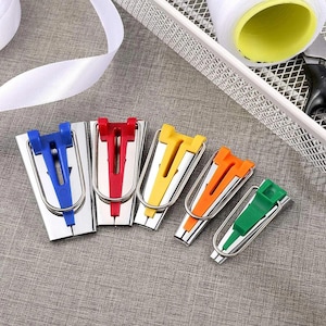May include: A set of five fabric bias tape makers in graduated sizes, featuring metallic silver bodies with colorful plastic inserts in blue, red, yellow, orange, and green. Each tool includes a wire handle for guiding fabric through the device.