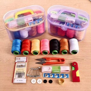 May include: A sewing kit featuring two clear plastic storage cases with handles, filled with assorted colorful thread spools, a pack of needles, safety pins, a measuring tape, a seam ripper, metal buttons, and a gold-tone metal thimble.