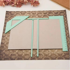 Könnte beinhalten: Ein Set aus mintgrünen Kunststoff-Buchbindewerkzeugen, bestehend aus einem Eckanleger, einem T-Lineal und einem Maßstab, platziert auf einem rechteckigen grauen Karton, der auf einem dunkelbraunen Papier mit goldenem Blumenmuster liegt.