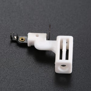 May include: A white plastic sewing machine part featuring a rectangular frame with a central vertical bar and a circular opening. Attached to the side is a black metal component with a small brass-colored eyelet and two thin metal prongs.
