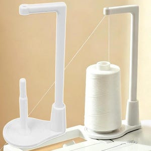 May include: A white plastic thread stand featuring a vertical arm with a top guide and a base pin for holding a large spool of thread. The stand is designed to support a spool of white thread, which is shown positioned on the base of the stand.
