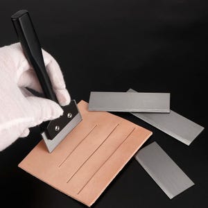 Könnte beinhalten: Ein Lederbearbeitungswerkzeug mit schwarzem Griff und silberner Metallklinge, das gerade einen geraden Schnitt in ein quadratisches Stück hellbraunes Leder macht. Drei zusätzliche rechteckige silberne Ersatzklingen liegen daneben.