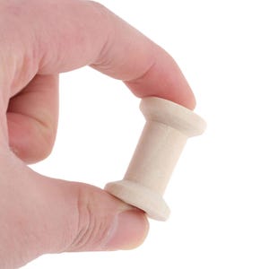 May include: A small, unfinished light-colored wooden spool with a cylindrical center and two flared circular ends. The spool features a smooth, natural wood grain texture and is designed for winding thread, yarn, or ribbon for various craft projects.