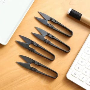 Puede incluir: Cuatro cortahilos de metal negro mate dispuestos en diagonal. Cada herramienta cuenta con una hoja puntiaguda, un mecanismo de bisagra con resorte y un diseño de mango de bucle continuo. Están fabricados en una sola pieza de metal con tornillos.