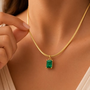 May include: A gold-colored necklace with a rectangular emerald-green pendant. The pendant is set in a gold frame and hangs from a delicate gold chain. The necklace is displayed on a person's neck.