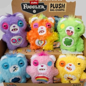 May include: A Zuru Fuggler plush bag charm display box containing six colourful, fuzzy bear-like creatures. Each plush features human-like teeth, mismatched eyes, and unique belly patches including a lollipop, rainbow heart, clover, cloud, rainbow, and sun.