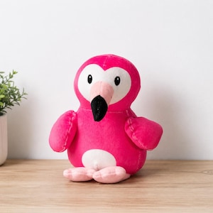 May include: A plush flamingo toy featuring a bright pink body, a white heart-shaped face patch, and a light pink beak with a black tip. The stuffed animal has small black eyes, two rounded wings, and two small light pink feet at the base.