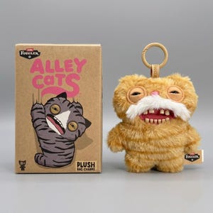 May include: A Fuggler Alley Cats plush bag charm with orange fur, a white moustache, and realistic teeth, attached to a plastic ring. Beside it is a brown cardboard box featuring the text Fuggler Alley Cats and an illustration of a grey cat.