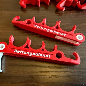 Può includere: Clip in plastica rossa con la scritta "Rettungsdienst" stampata in bianco. Le clip hanno un design unico con più sezioni curve e una cerniera bianca. Diverse clip sono visibili su una superficie di legno scuro.