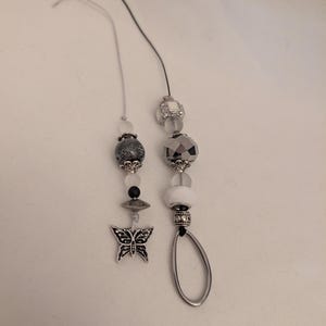 May include: Two beaded charms with silver and white beads. One charm features a butterfly pendant. The other charm has a silver oval-shaped pendant. Both charms have black and gray string.