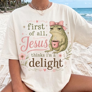 May include: An off-white short-sleeve t-shirt featuring a graphic of a green toad wearing a pink bow and holding a pink mug. The text reads: first of all, Jesus thinks i'm a delight. The design includes pink hearts, small stars, and floral accents.