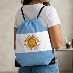 May include: A blue, white, and gold drawstring backpack featuring the flag of Argentina. The backpack has black drawstrings and black reinforced corners. The Argentinian flag design includes a sun with a face in the center.