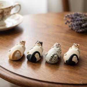Könnte beinhalten: Vier Keramik-Katzenfiguren in entspannter Pose, jede mit einem einzigartigen Farbmuster. Die Katzen liegen auf dem Rücken, die Pfoten in der Luft. Die Figuren stehen auf einem Holztisch, im Hintergrund eine Tasse und Untertasse.