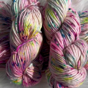 May include: Two skeins of thick, soft yarn featuring a pale pink base color with vibrant speckles of bright yellow, neon green, magenta, and deep blue. The yarn is twisted into a traditional hank shape, showcasing a multi-colored, speckled appearance.