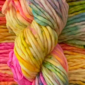 May include: A thick, bulky skein of yarn featuring a vibrant, multi-colored hand-dyed pattern. The yarn displays a blend of bright pink, sunny yellow, soft orange, and light blue hues, twisted into a dense, soft, and textured fiber coil.