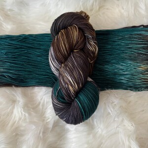 May include: A skein of hand-dyed yarn in shades of brown, gray, white, and teal. The yarn is arranged in a twisted bundle, with a length of teal yarn extending to the side. The background is a soft, white, fluffy material.