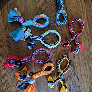 May include: A collection of colorful dog toys made from braided fabric. The toys feature loops and knotted ends with various color combinations, including blue, red, orange, and yellow. They are arranged on a wooden table.