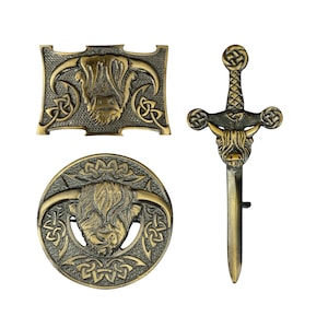 May include: A set of three antique brass-toned metal accessories featuring Highland cow motifs and Celtic knotwork. The collection includes a rectangular belt buckle, a circular brooch, and a sword-shaped kilt pin with a cow head detail.