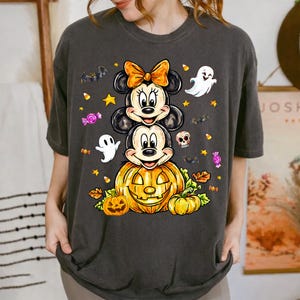 Könnte beinhalten: Ein anthrazitfarbenes T-Shirt mit einer Grafik von Minnie und Mickey Mouse, die vertikal gestapelt sind, wobei Minnie eine orangefarbene Schleife trägt. Darunter befindet sich ein großer Kürbis, umgeben von kleinen Geistern und Fledermäusen.