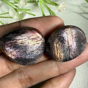 May include: Two polished sunstone gemstones. The stones have a dark base color with shimmering inclusions of pink, gold, and silver. The stones are oval and teardrop shaped. The gemstones are held in a hand.