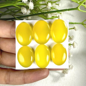 May include: Six oval-shaped, polished yellow gemstones arranged on a white card. The gemstones have a smooth, glossy surface and are a vibrant, sunny yellow color. The background includes white flowers and green stems.