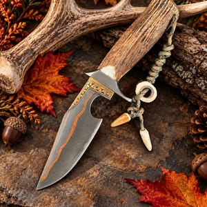 May include: A fixed-blade knife with a textured antler handle and a dark, patterned steel blade featuring a prominent orange copper-colored inlay. The knife includes a metal bolster with intricate engravings and a lanyard with small bone beads.