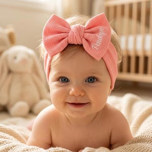 Pode incluir: Um bebê usando uma faixa de cabelo coral com um laço grande. A faixa é bordada com o nome "Penelope". O bebê está sorrindo e tem olhos azuis. Um coelho de pelúcia e um berço estão ao fundo.