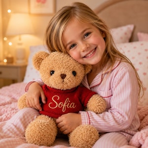 Könnte beinhalten: Ein hellbrauner Teddybär mit einem roten Pullover, auf dem der Name "Sofia" gestickt ist. Der Bär wird von einem Kind umarmt, das einen rosa gestreiften Pyjama trägt. Der Teddybär hat ein strukturiertes Aussehen.