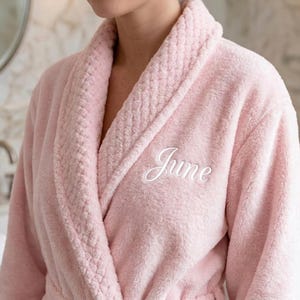 以下が含まれることがあります： 胸に白い筆記体で「June」と刺繍された、柔らかく淡いピンク色の豪華なバスローブ。 ショールカラーと、襟と袖口にテクスチャード加工が施されており、居心地が良く贅沢な雰囲気を醸し出しています。