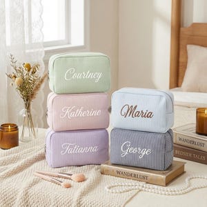 Könnte beinhalten: Sechs personalisierte Kosmetiktaschen in Pastellfarben: Grün, Rosa, Lavendel, Hellblau und Marineblau. Jede Tasche hat einen Namen in einer komplementären Farbe gestickt. Die Taschen sind auf einem Tisch gestapelt.