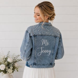 Puede incluir: Una chaqueta de mezclilla azul de tono medio con cuello, mangas largas y cierre de botones. El panel superior de la espalda está adornado con numerosas perlas blancas. El texto "Ms. Jenny" está impreso en letra cursiva blanca en la espalda.