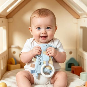 May include: A smiling baby holds a blue and yellow plush bunny toy with a white teething ring. The toy is labelled "Daria". The baby is wearing a white onesie. The toy is designed for babies.