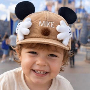 Puede incluir: Gorra de béisbol de pana color beige con orejas de ratón negras, manos enguantadas blancas y el nombre "MIKE" bordado en blanco. Un pompón marrón está centrado en la gorra. La gorra la lleva un niño sonriente.