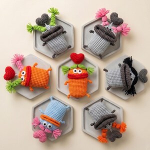 Puede incluir: Una colección de siete figuras de monstruos de ganchillo hechas a mano en varios colores, incluyendo naranja, azul y gris. Cada monstruo tiene características únicas, como acentos en forma de corazón y pelo colorido, y se muestra en una bandeja hexagonal gris.