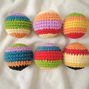 May include: Six colorful, handmade crochet balls. Each ball features horizontal stripes in various colors, including red, orange, yellow, green, blue, and purple. The balls are arranged on a white fabric background.